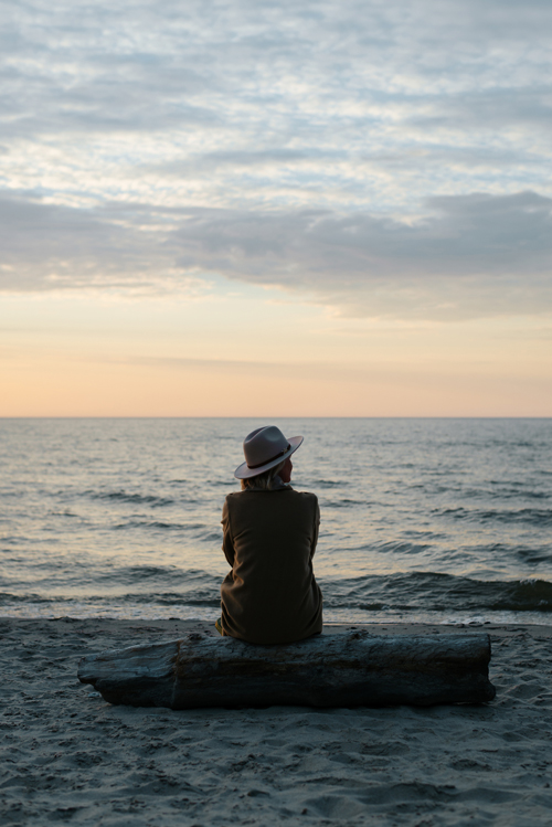 Photo of back of Kelly Taylor sitting on log reflecting on beach